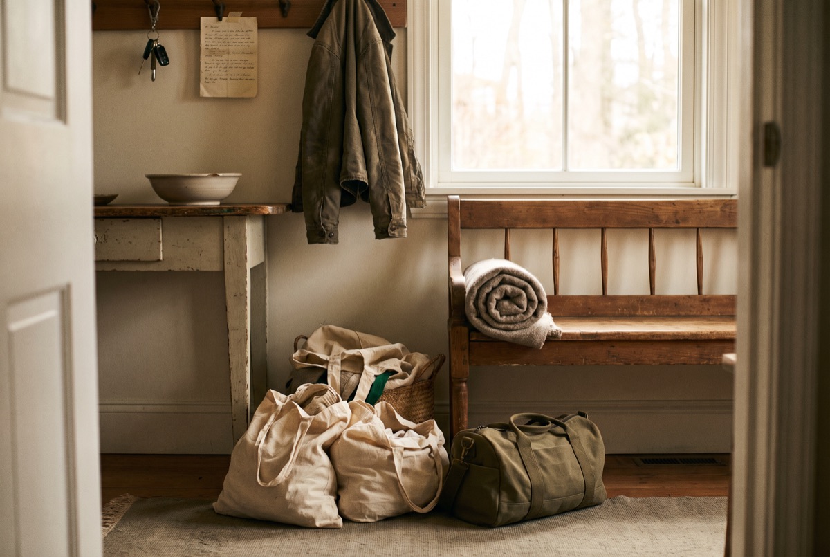A farmhouse front hall with packed canvas totes and an olive duffel staged on the rug, a rolled blanket on a wooden bench, keys and a waxed jacket hanging by the door.